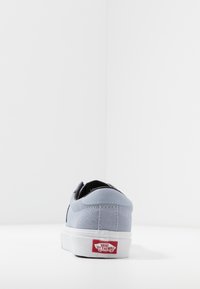 Zapatilla baja de ante gris con suela de goma blanca. Presenta un forro negro y una etiqueta con logo rojo en el talón. Textura suave y forma redondeada.