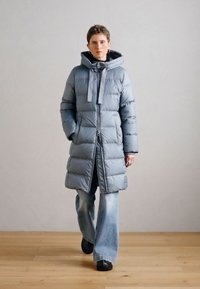 Lichtblauwe pufferjas met een lange silhouette, ritssluiting en verstelbare capuchon. Voorzien van horizontale doorgestikte secties en zijzakken.