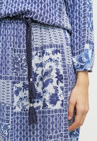 Haut en tissu à motifs bleus avec un cordon de serrage et un détail de pampille à la taille, présentant des designs floraux et géométriques dans différentes nuances de bleu.