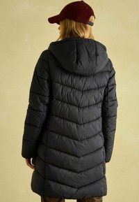 Melna pūkaina jaka ar zigzag quiltēšanas rakstu un kāju. Valkāta ar burgundijas krāsas cepuri, kas izceļ gludu un spīdīgu tekstūru.