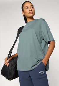 Mehakko zelena majica z dolgimi rokavi iz bombaža, v velikosti oversized, v kombinaciji z mornarsko modrimi hlačami z belim logotipom, skupaj z črno športno torbo.