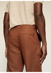 Un pantalon en lin marron présente une coupe ajustée, deux poches arrière avec des boutons et une texture lisse. Porté avec un haut en tissu beige.