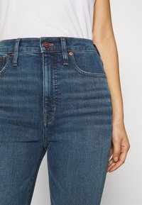 Person som bär högmidjade blå denimjeans med kopparnitar och en vit skjorta instoppad, med handen vilande vid sidan.