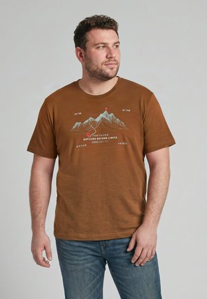Mężczyzna z krótkimi kręconymi włosami, ubrany w brązowy t-shirt z motywem górskim i niebieskie jeansy, stoi na jednolitym jasnym tle.