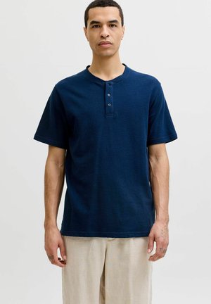 Homme debout portant un t-shirt henley bleu marine à manches courtes avec trois boutons et un pantalon beige clair sur un fond uni.