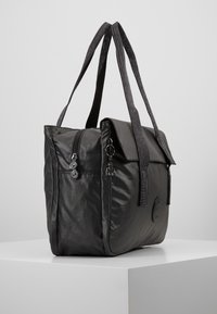 Sac cabas en nylon noir avec deux anses texturées, fermeture éclair, rabat pliable et détail logo sur le côté. Texture lisse et brillante.