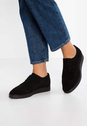 Scarpe oxford in suede nero con punta arrotondata, dettagli testurizzati e suola nera bassa, indossate con jeans blu denim risvoltati.