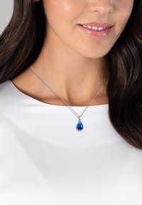 Silberne Halskette mit einem tropfenförmigen blauen Edelsteinanhänger, getragen auf einem weißen Oberteil. Verfügt über eine zarte Kette und eine polierte Oberfläche.