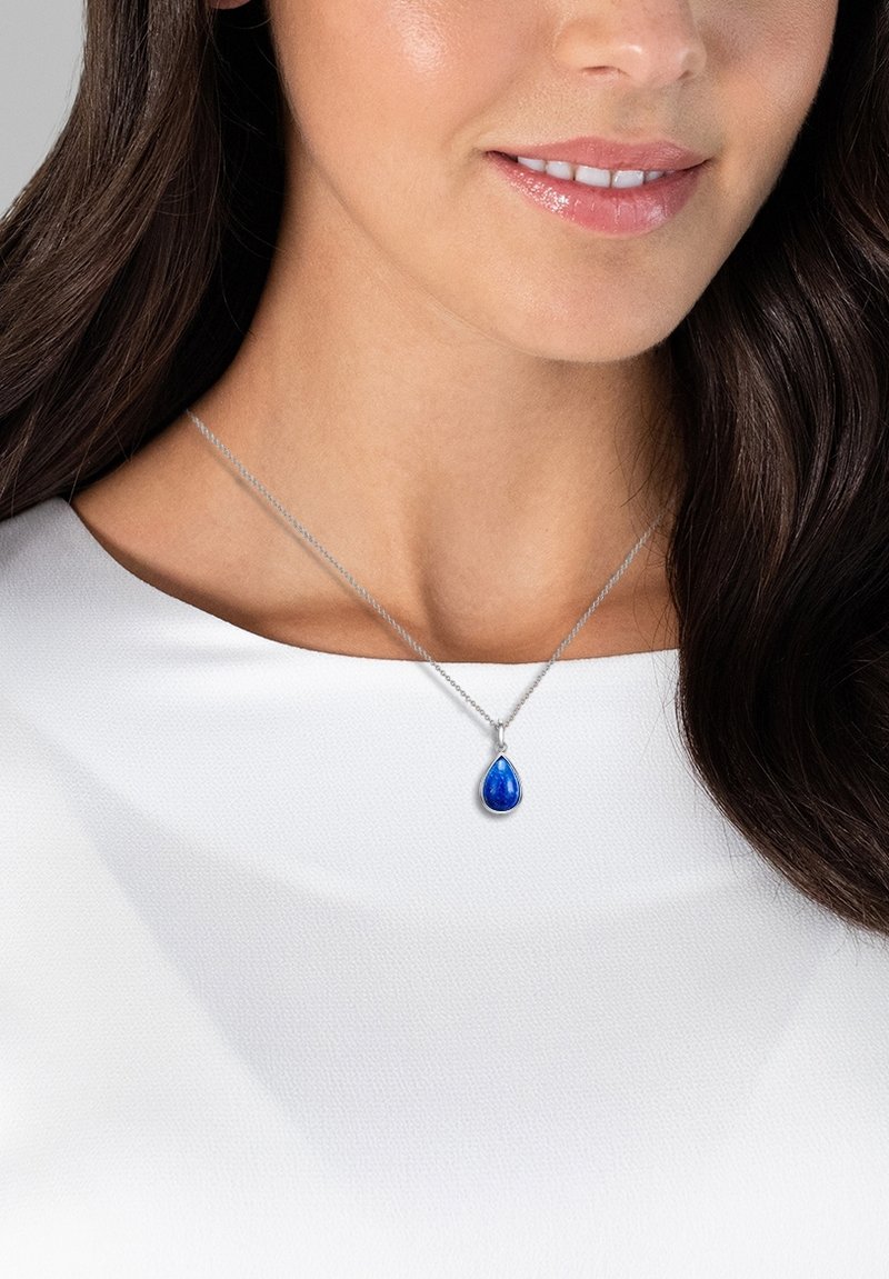 Silberne Halskette mit einem tropfenförmigen blauen Edelsteinanhänger, getragen auf einem weißen Oberteil. Verfügt über eine zarte Kette und eine polierte Oberfläche.