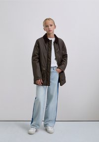 Giacca imbottita marrone con colletto scuro, t-shirt bianca, jeans azzurri chiari con strisce laterali e sneakers bianche. Design semplice, vestibilità comoda.