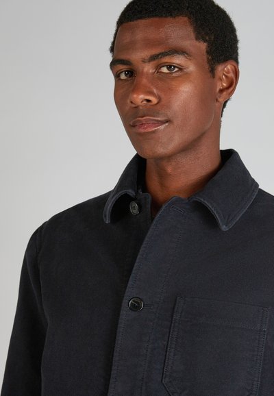 Mężczyzna o ciemnej skórze i krótkich kręconych włosach, ubrany w ciemną kurtkę z guzikami, z kołnierzem i kieszenią na piersi, zwrócony lekko w prawo.