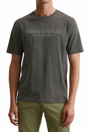 Mann trägt dunkelgraues T-Shirt mit kurzen Ärmeln und dem Schriftzug „Marc O’Polo Est. in Stockholm“ und olivgrüne Hose und steht vor weißem Hintergrund.