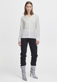 Camicia bianca a maniche lunghe con bottoni realizzata in un tessuto leggero, abbinata a pantaloni neri a vita alta e stivali alti d'argento con tacco.