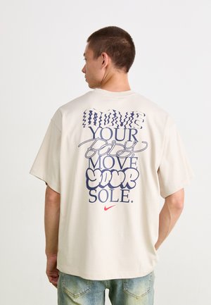 T-shirt oversize beige clair avec un texte navy audacieux et un logo Nike rouge au dos. Manches courtes et texture de tissu douce.