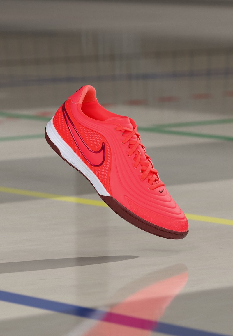 Klar rødt sportssko med svart Nike-swoosh og hvit såle som svever over gulvet på en innendørsbane med fargede linjer.