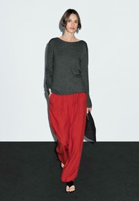 Une femme aux cheveux courts marche avec confiance dans un haut gris et un pantalon large rouge, tenant un sac noir, sur un fond clair.
