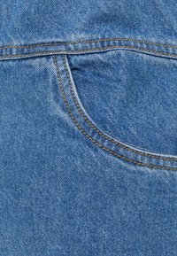 Jeansstof i ljusblått med synlig struktur och sömmar. Har en rundad jeansficka med kontrasterande gyllenbrun tråd.