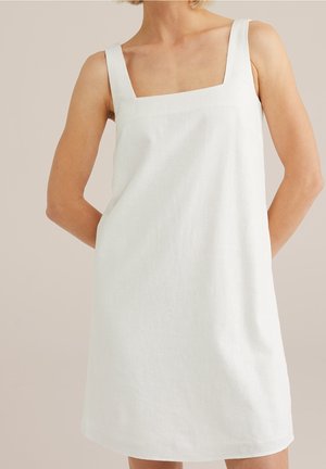 Femme portant une robe blanche sans manches avec un décolleté carré, debout les mains derrière le dos contre un fond uni.