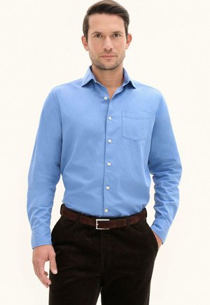 Mann trägt ein hellblaues Hemd mit Brusttasche, dunkelbraunem Gürtel und dunkler Kordhose, steht mit einer Hand in der Tasche.