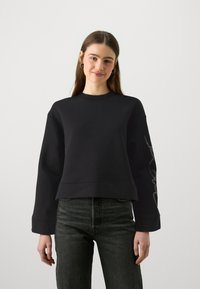 Zwarte cropped sweatshirt van zachte stof met een ronde hals en lange mouwen. Heeft een decoratief zilveren ontwerp langs de linkermouw.