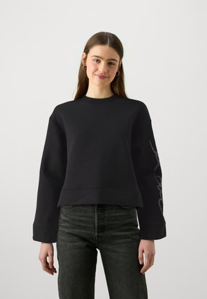 Zwarte cropped sweatshirt van zachte stof met een ronde hals en lange mouwen. Heeft een decoratief zilveren ontwerp langs de linkermouw.