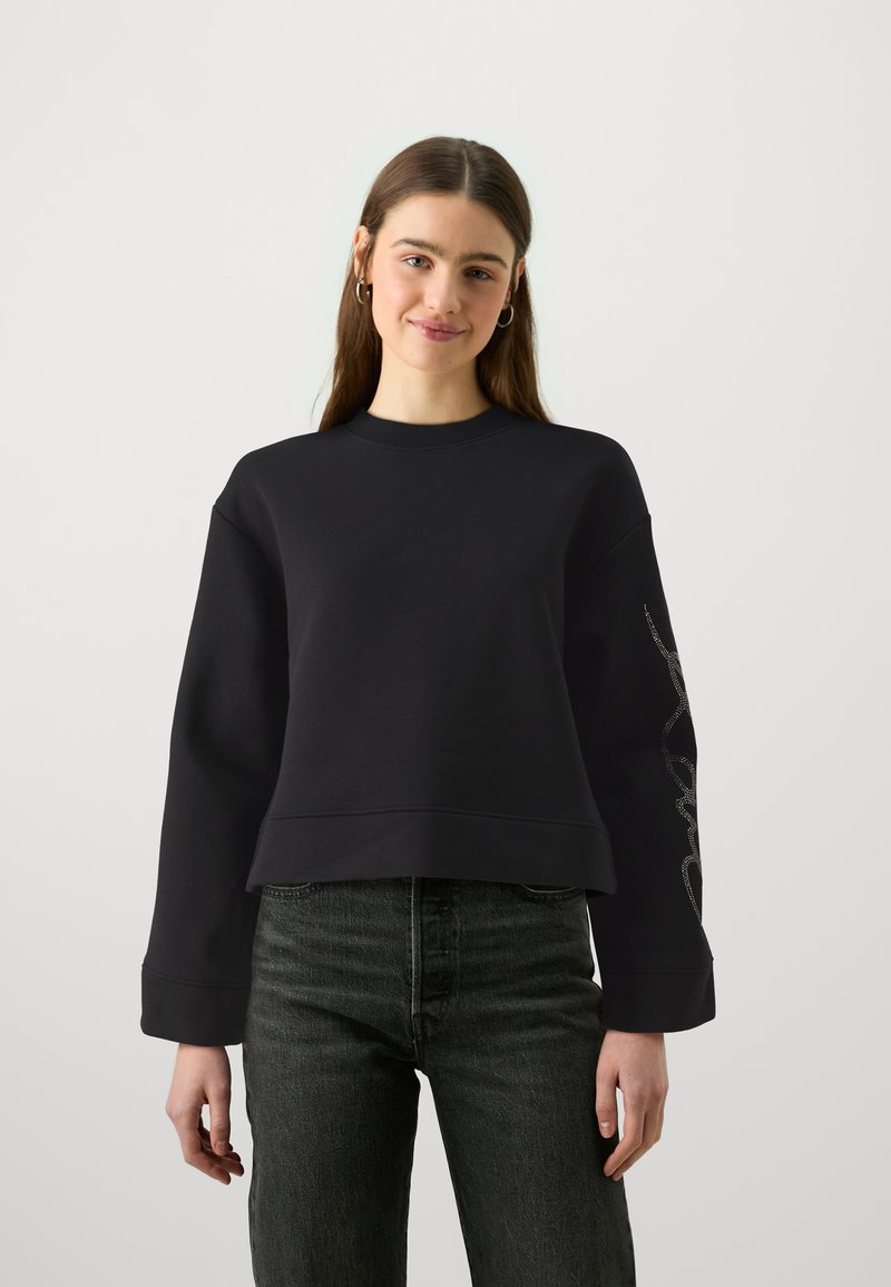 Zwarte cropped sweatshirt van zachte stof met een ronde hals en lange mouwen. Heeft een decoratief zilveren ontwerp langs de linkermouw.