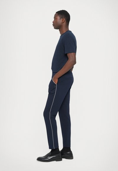 Navy Kurzarm-T-Shirt, kombiniert mit eng anliegenden navyfarbenen Hosen mit weißen Seitenstreifen. Die Schuhe sind schwarz mit glänzender Oberfläche.