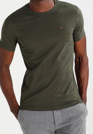 Uomo che indossa una maglietta a maniche corte aderente verde oliva con un piccolo logo Tommy Hilfiger e pantaloni grigio chiaro, con le mani in tasca.