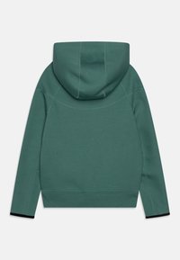 Zadní pohled na jednobarevnou zelenou mikinu s velkou kapucí, dlouhými rukávy a černými manžetami na bílém pozadí.