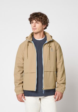Homme portant une veste zippée beige texturée avec capuche, un sweat-shirt bleu marine et un pantalon clair, debout devant un fond uni clair.