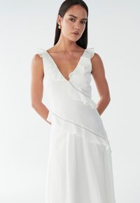 Weißes Kleid mit Rüschendetails entlang des V-Ausschnitts und der Seite; aus leichtem Stoff mit strukturierter Oberfläche und subtilen Nähdetails gefertigt.