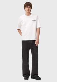 Valge puuvillane T-särk lühikeste varrukatega, ees logo ja mustad sirge jalaga teksad, seljas mustad kingad, tasasel taustal.