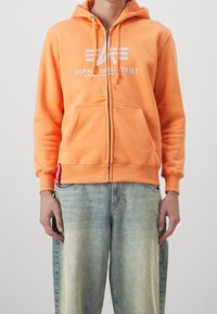 Felpa arancione con zip realizzata in cotone, caratterizzata da un logo bianco sul petto, due tasche frontali e polsini a costine. Abbinata a jeans azzurri chiari.