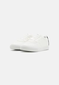 Hvite sneakers med teksturert overflate, glatt lærdesign, rund tå og kontrast svart hælaccent. Skoene har snøring og perforerte detaljer.