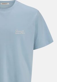 Camiseta de algodón azul claro con mangas cortas, cuello redondo y un pequeño logotipo bordado en blanco en el pecho. Textura suave.