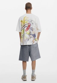 Tricou alb supradimensionat cu un design colorat cu tucan și palme pe spate. Asortat cu pantaloni scurți până la genunchi, în dungi albastre și albe.