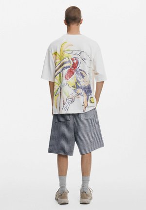 Camiseta blanca de gran tamaño con un diseño colorido de tucán y palmera en la parte posterior. Combinada con shorts hasta la rodilla a rayas azules y blancas.