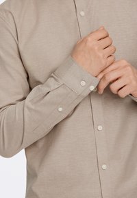 Μπεζ μακρυμάνικο πουκάμισο με σχεδιασμό με κουμπιά, διακριτή υφή και κανονικά μανίκια με λευκά κουμπιά. Το ύφασμα Jockey πέφτει απαλά.