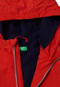 Rote Jacke mit Kapuze, ausgestattet mit einem marineblauen Fleecefutter, einem silbernen Reißverschluss und einem grünen Etikett im Inneren. Das Außenmaterial ist glatt und wasserabweisend.