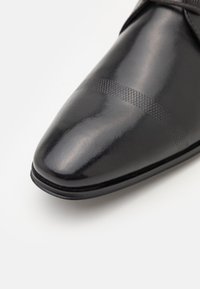 Chaussure en cuir noir avec un bout pointu, présentant un dessus lisse et un motif rayé diagonal texturé au centre.