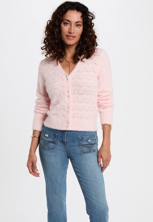 Lichtroze gebreide cardigan met een v-hals, een gestructureerd patroon en knopen. Gecombineerd met blauwe jeans met decoratieve voork pocketen.