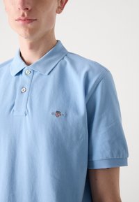Lyseblå polo skjorte laget av bomull med en teksturert overflate. Har en tradisjonell krage, to knapper, og en liten brodert logo på brystet.