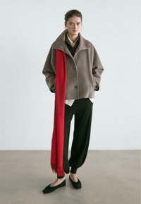 Brązowa kurtka z mieszanki wełny z szerokim kołnierzem i guzikami, zestawiona z długim czerwonym szalem. Czarne, luźne spodnie i czarne płaskie buty.