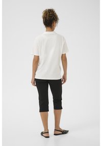 Witte katoenen polo met kraag, gecombineerd met zwarte capri-broekjes en zwarte sandalen. Van achteren gefotografeerd, waardoor de eenvoud van de outfit goed tot uiting komt.