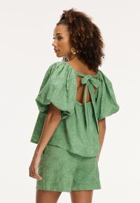 Blusa verde in cotone con maniche a sbuffo, fiocco sul retro e motivi floreali a rilievo. Abbinata a pantaloni corti coordinati, anch'essi a rilievo.