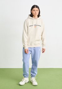 Hanorac crem cu logo-ul "New Balance" în negru și portocaliu, asortat cu pantaloni de trening bleu deschis și adidași albi cu tălpi texturate.