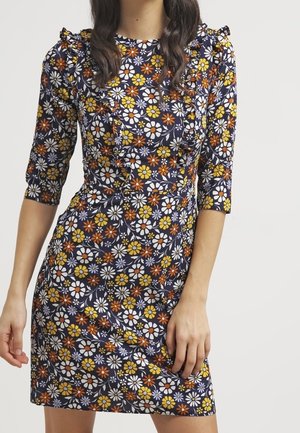 Vrouw draagt een aansluitende marineblauwe jurk met oranje, gele en witte bloemenprint en ruches op de borst en schouders.