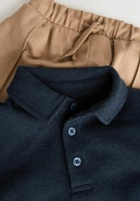 Marineblaues Poloshirt aus strukturiertem Stoff mit drei blauen Knöpfen und einem geschwungenen Kragen, kombiniert mit beigen Jogginghosen mit elastischem Bund.