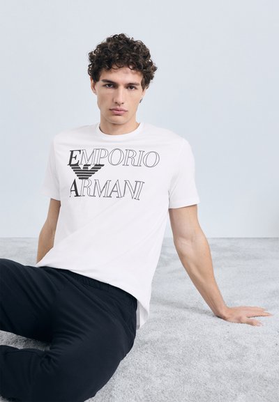 Wit katoenen T-shirt met de tekst "EMPORIO ARMANI" in zwarte letters. Model zittend op een grijze ondergrond, draagt zwarte elastische broek met tailleband.