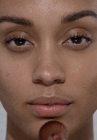Nahaufnahme eines Gesichts mit glatter Haut. Natürlicher Make-up-Look, sichtbare Wimpern und eine sanfte rosa Lippenfarbe. Ein Pinsel berührt das Kinn.